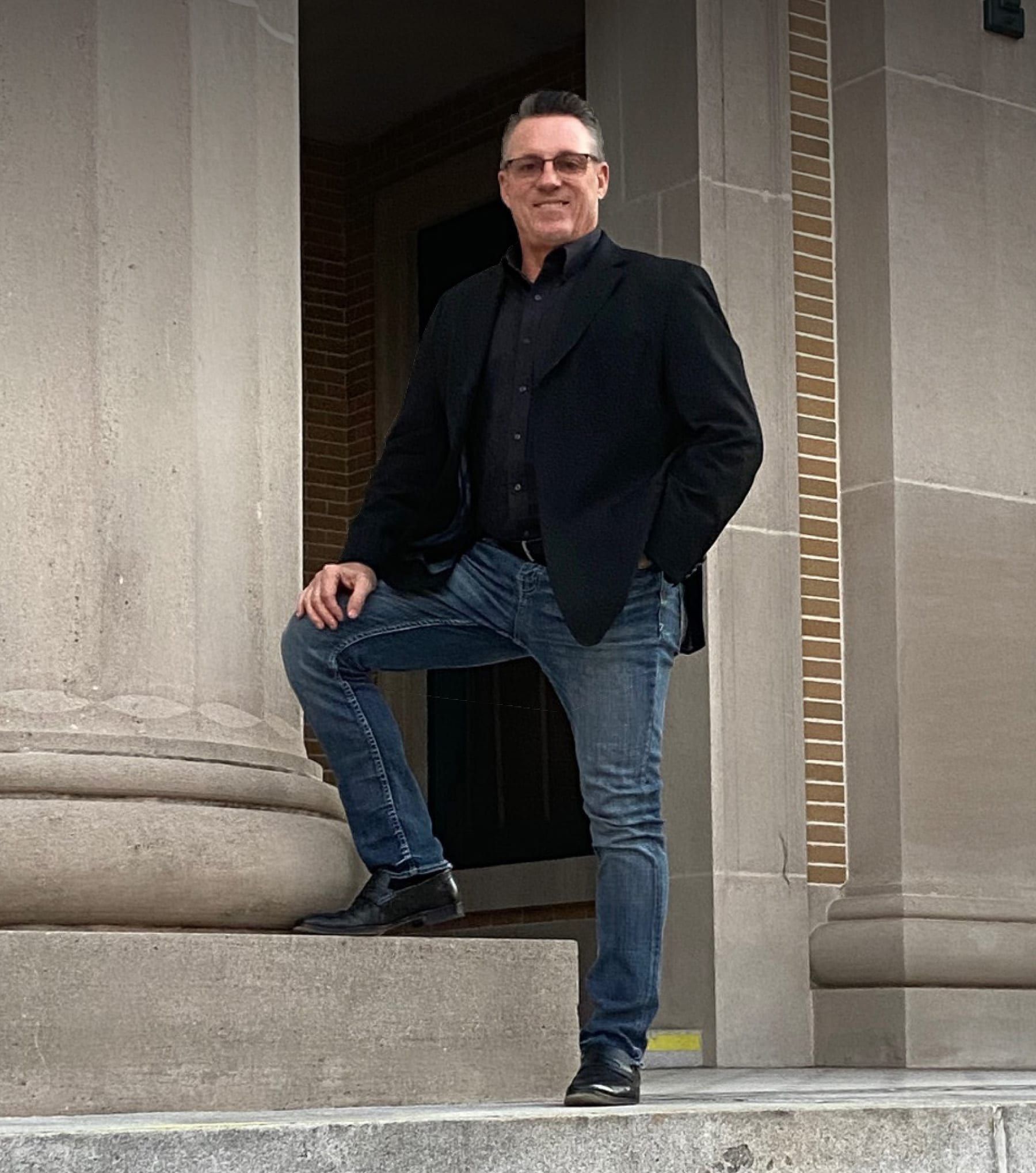 Man standing on stone steps by a large columned building, wearing a dark jacket and jeans.