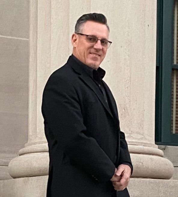 Man in a black coat and glasses standing by a stone column, smiling slightly.