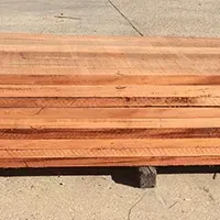 Stack of reddish-brown lumber planks, resting on a block in an outdoor setting.