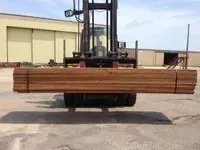 Forklift carrying a bundle of brown lumber in an outdoor setting. The building is visible in the background.