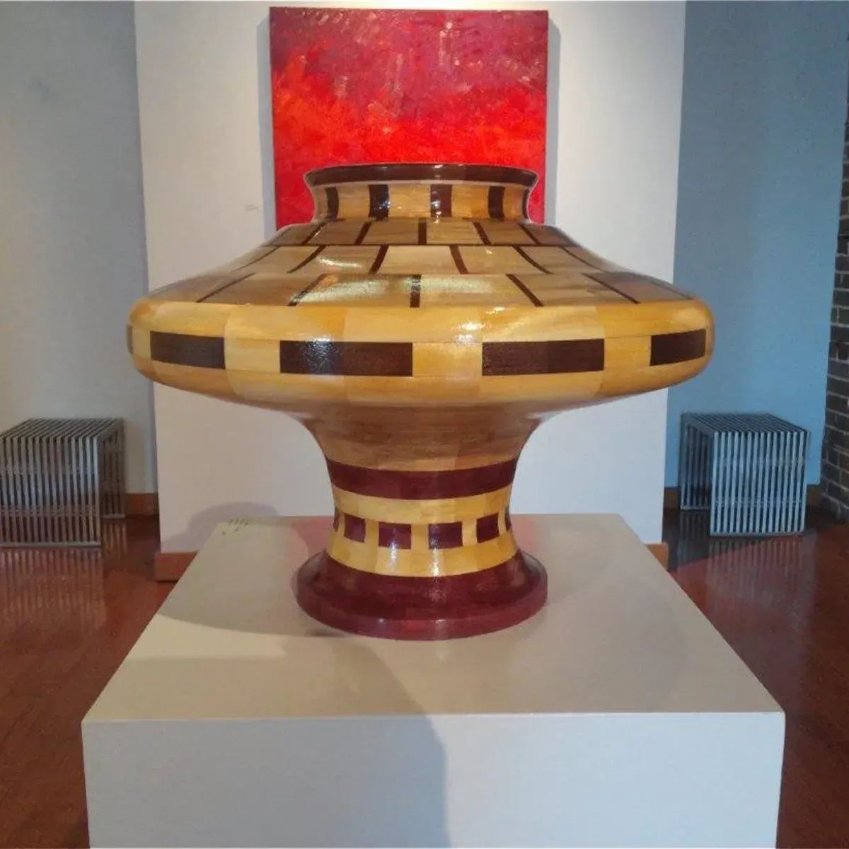 Large, ornate wooden vase with a tiered design, displayed on a white pedestal against a red and white background.