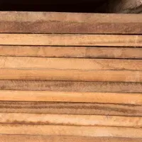 Stack of rough-cut, light-brown wooden boards, seen from the side, arranged horizontally.