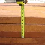 Tape measure showing the thickness of stacked, reddish-brown wooden planks. A hand holds the measuring tape.