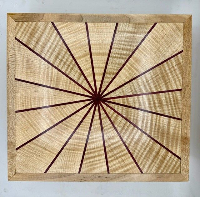 Wooden square with radial design: light wood with dark red lines radiating from a central point, framed by light wood.