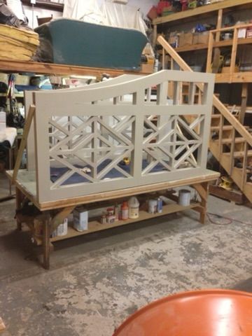 A partially constructed, light-colored wooden headboard with a geometric design sits on a workbench in a cluttered workshop.