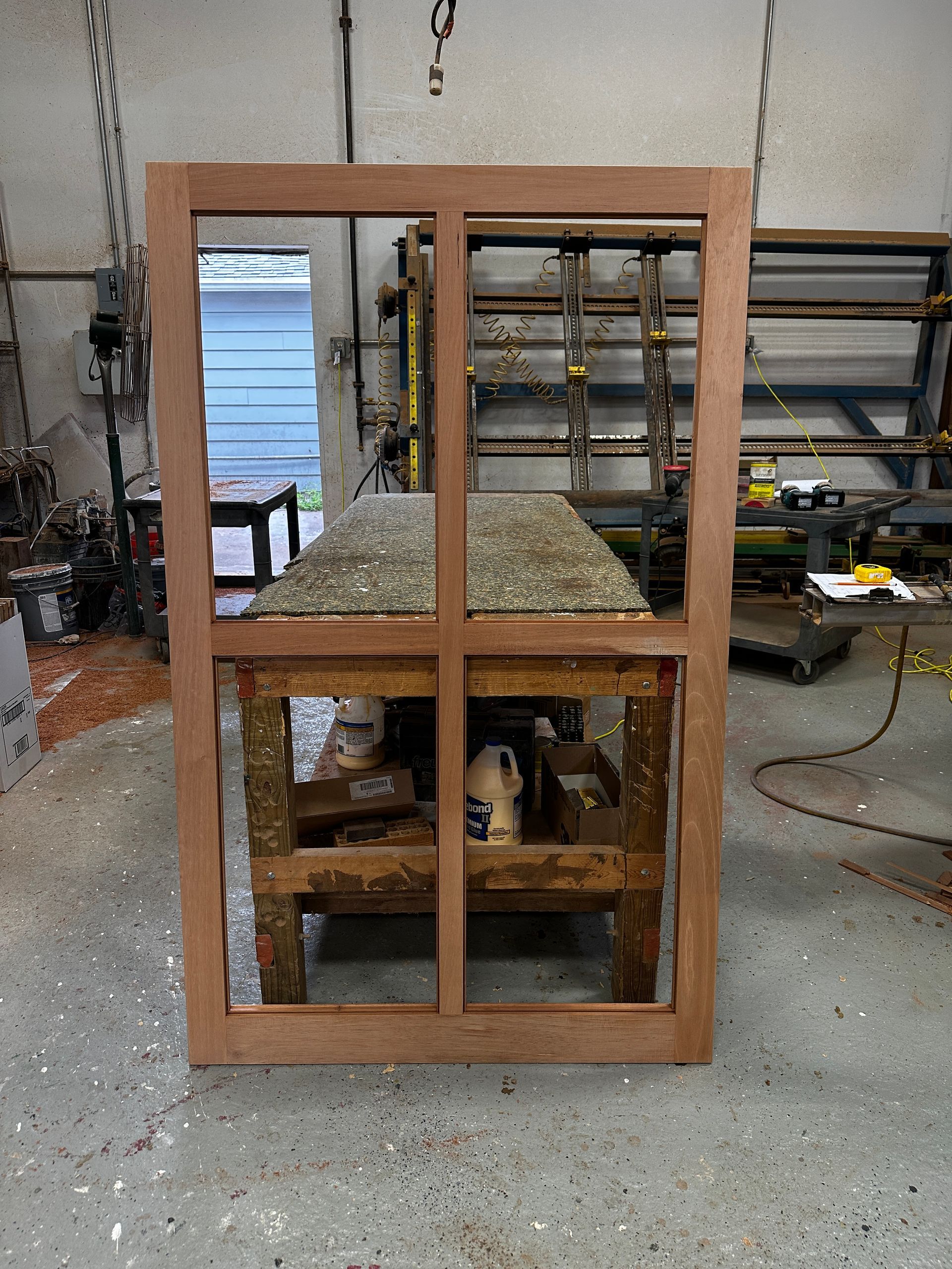 A wooden frame for a window, with two panes, sits on a workbench in a workshop. The frame is brown.