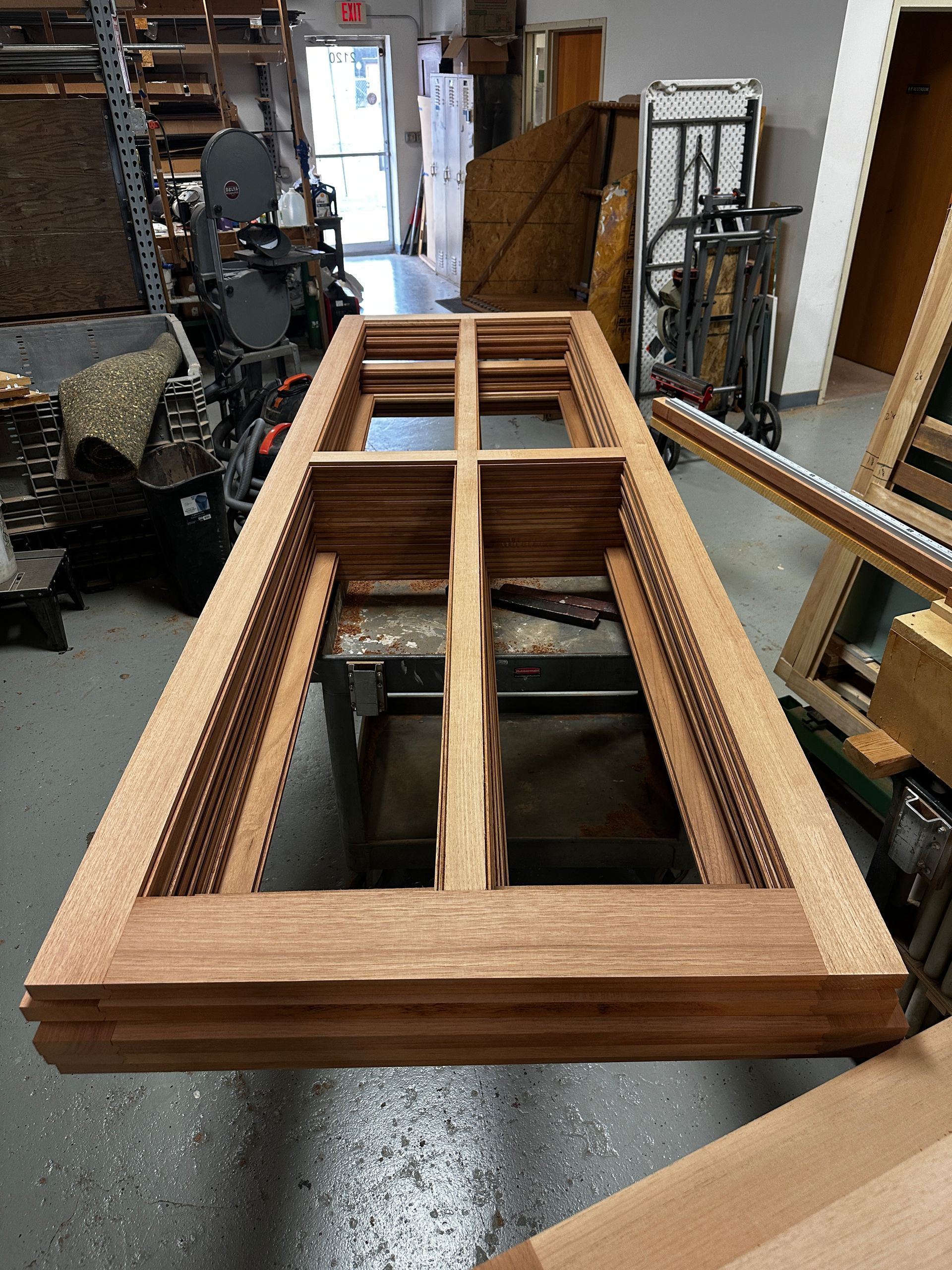 Wooden window frames stacked in a workshop, showing the grid pattern for glass panes. The frames are made of light brown wood.