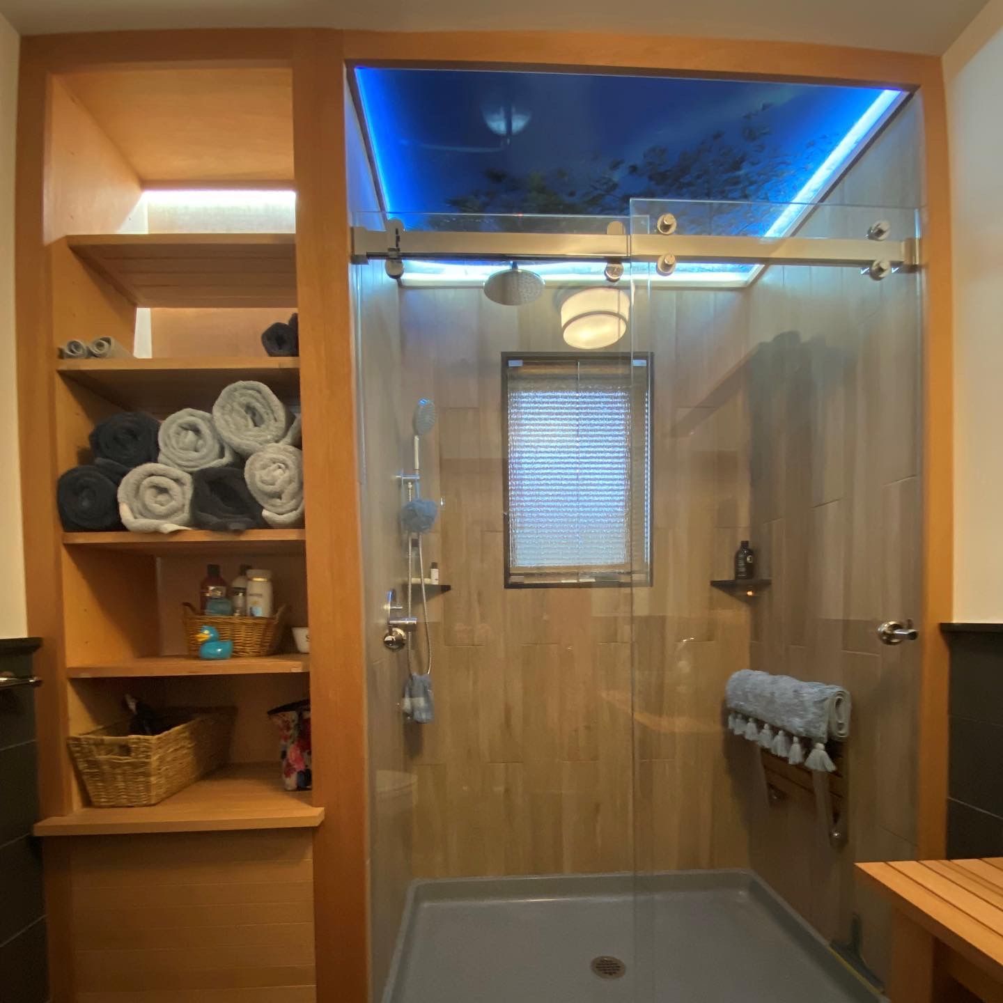 A modern bathroom with a wooden built-in shelf unit holding rolled towels and toiletries next to a glass shower with a gray floor.