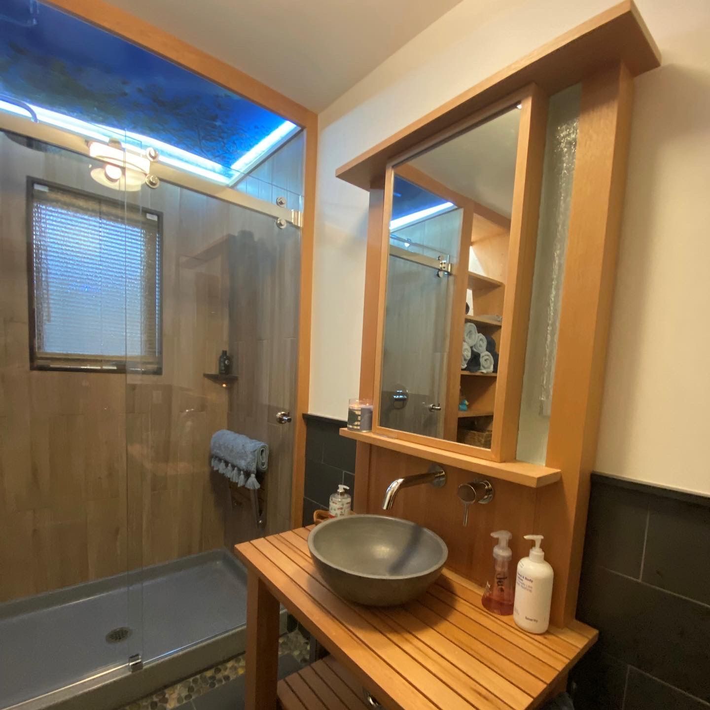 Bathroom with a wooden vanity, vessel sink, and large mirror. Shower stall on the left with natural light from above.