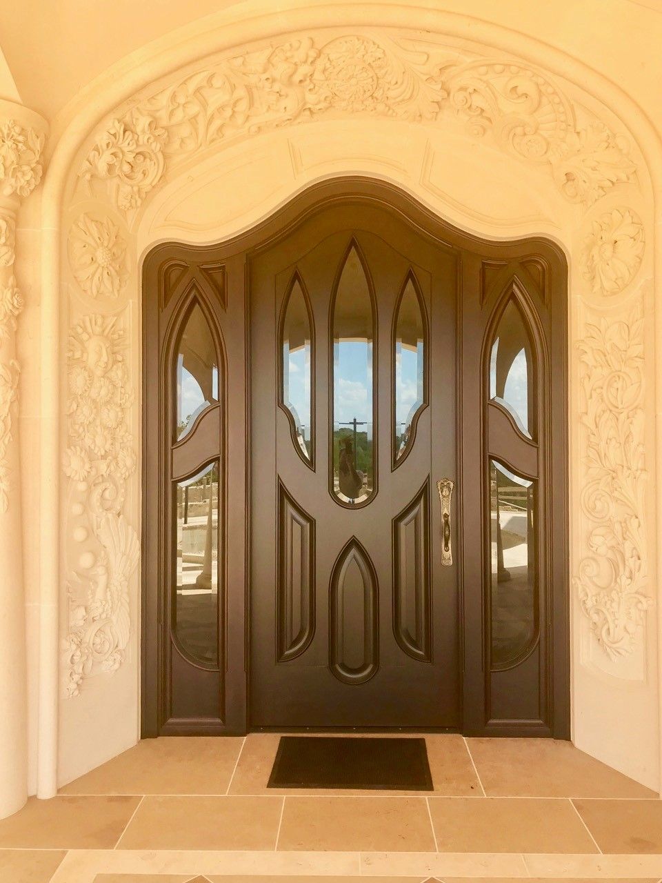 Ornate brown front door with glass panels set in a beige arched doorway with decorative relief, on a stone patio.