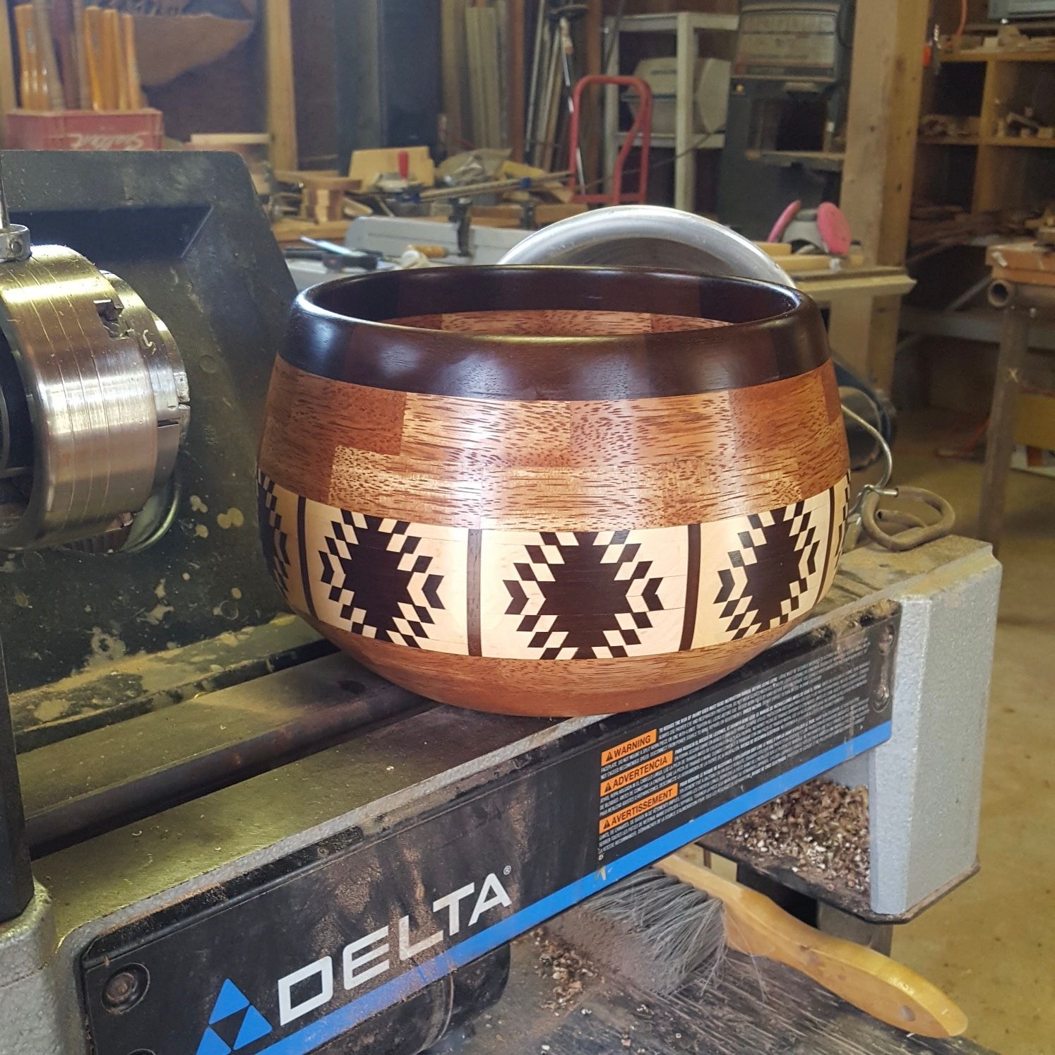 A wooden bowl with a geometric design on a wood lathe in a workshop. The bowl has a dark rim and light and dark wood bands.