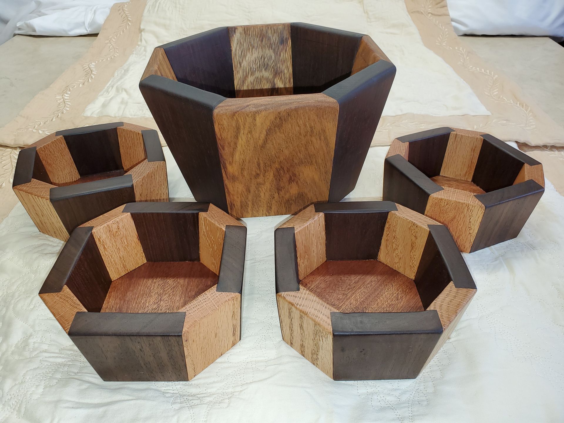 Wooden polygonal bowls in various sizes, arranged on a cream-colored surface. The bowls have alternating dark and light wood sections.