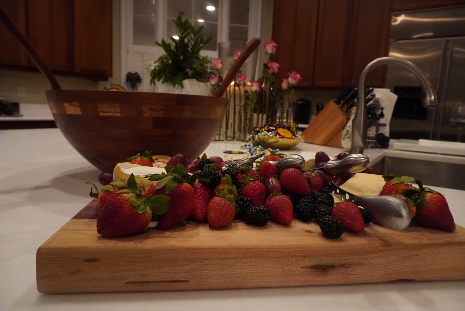 A wooden charcuterie board laden with fresh berries and cheese sits on a kitchen counter. A large salad bowl and a sink are in the background.