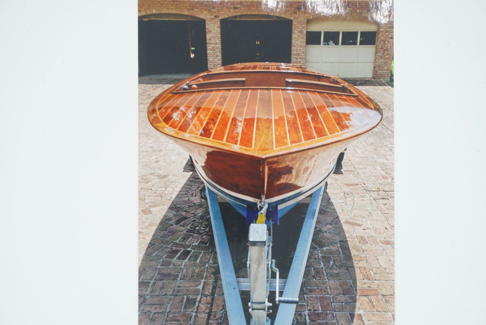 A wooden boat, with a glossy varnished deck, sits on a blue trailer in front of a brick building with garage doors.