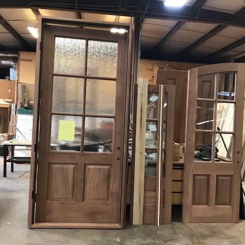 Wooden doors with glass panels are stacked in a workshop. Some have a traditional design, others are arched.
