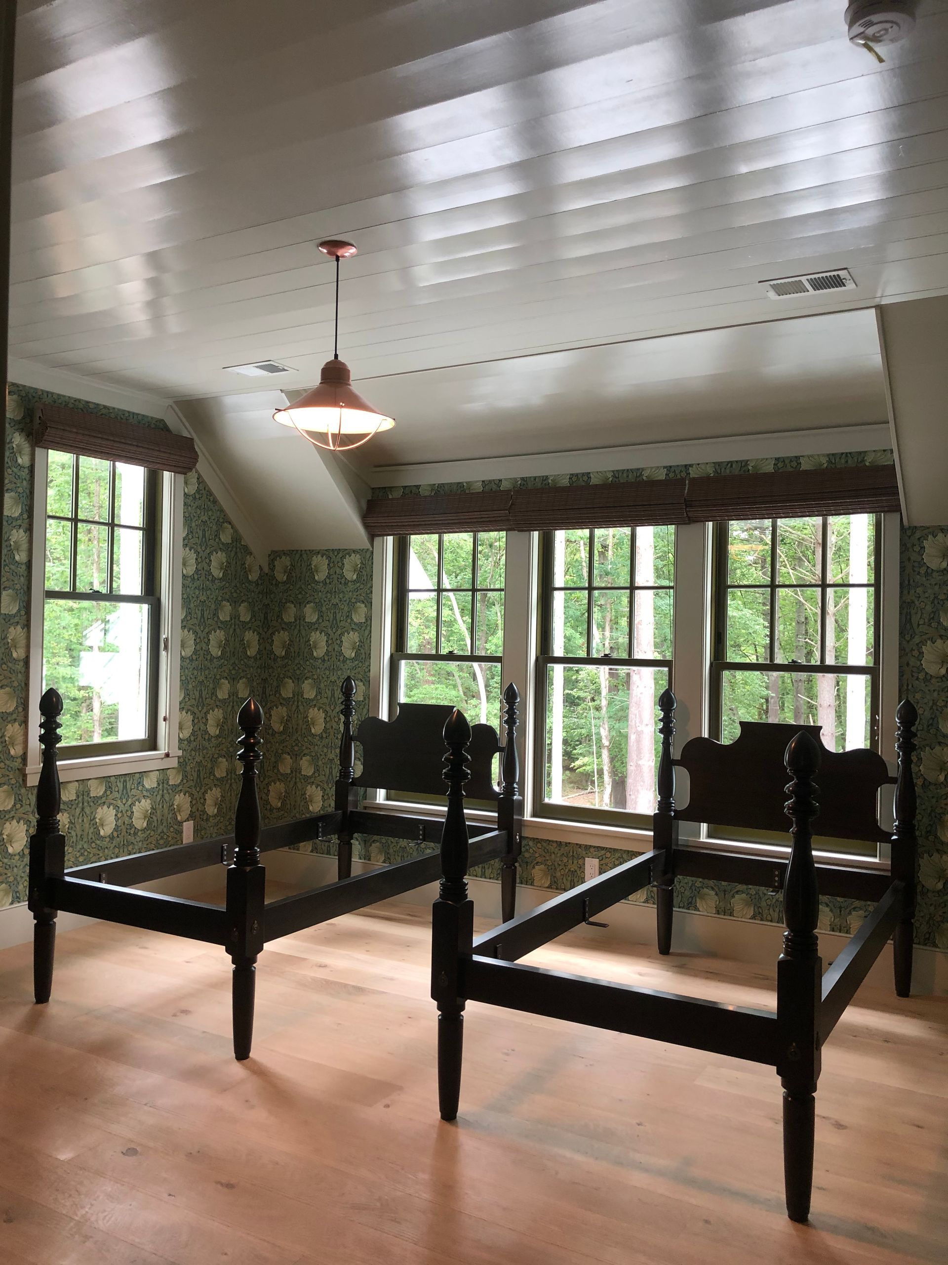 Two empty black bed frames in a bedroom with floral wallpaper, windows, and a hanging light.