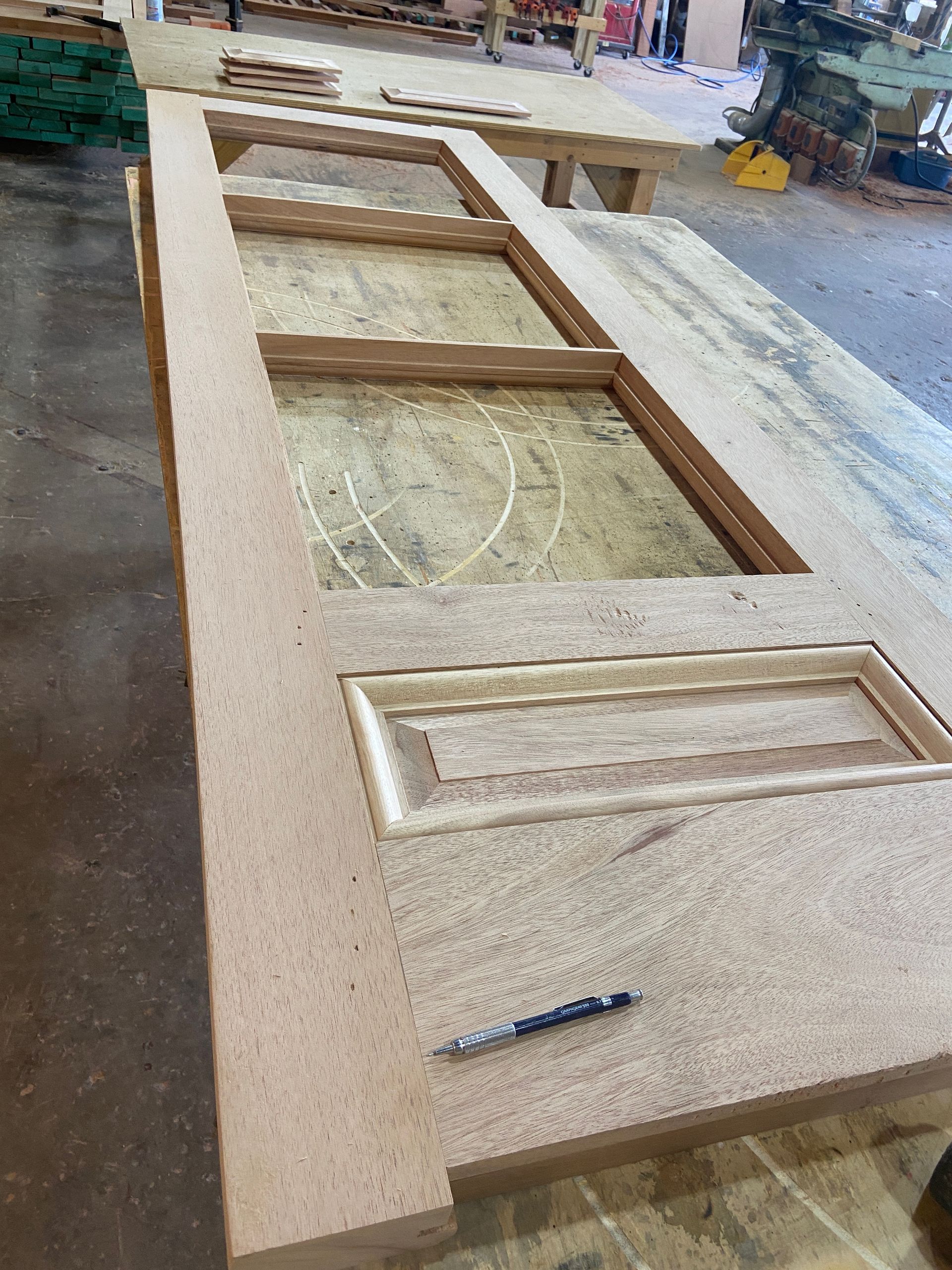 A wooden door frame under construction on a workbench, with sections carved out and a pencil lying on the surface.