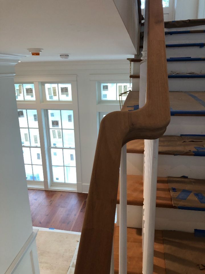 Wooden banister with a curved, decorative detail on a staircase.  Stair treads are covered with protective blue tape, white walls, and large windows.