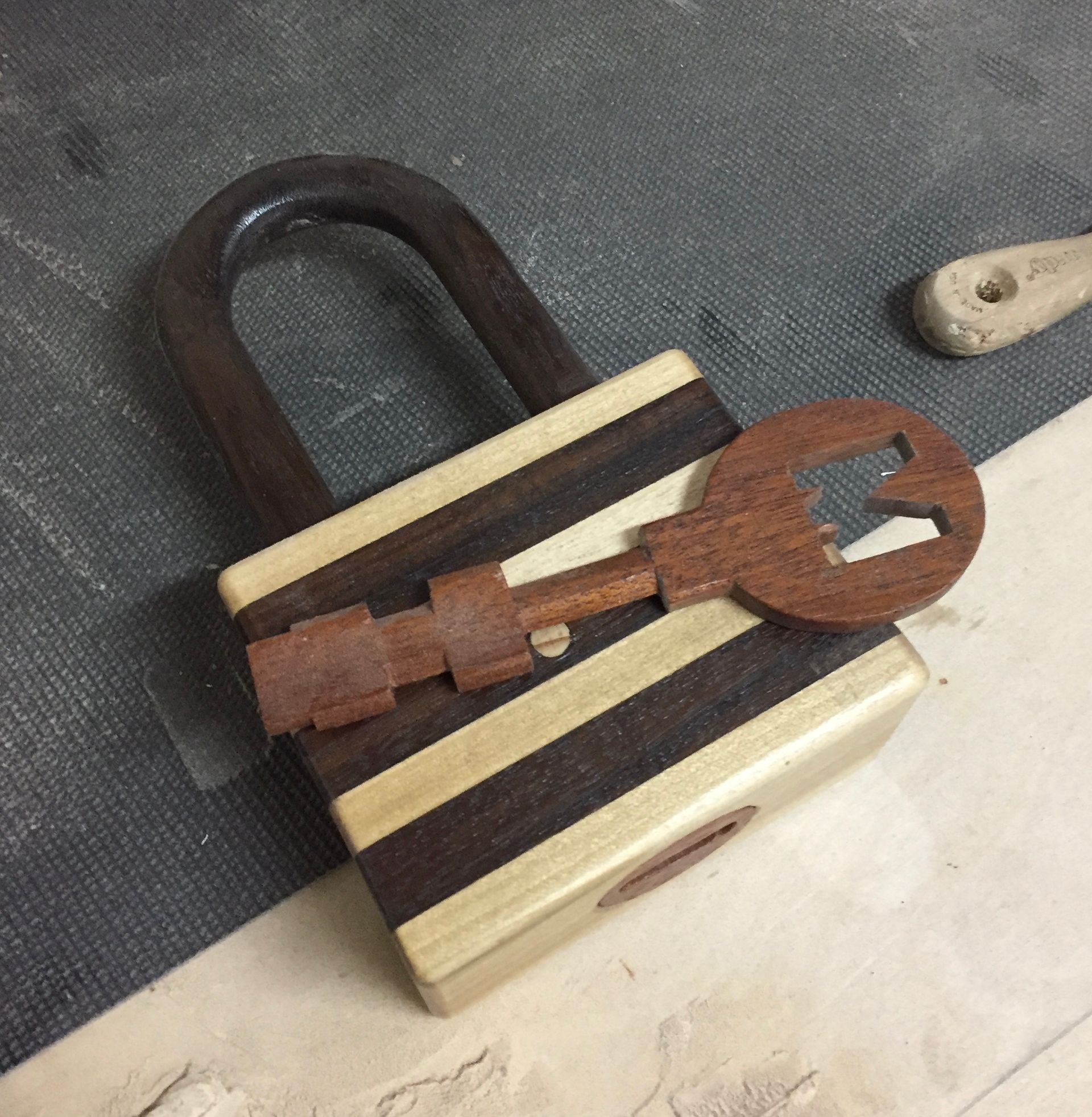 Wooden padlock with a matching key lying on top of it, against a textured surface. The lock is made of alternating light and dark wood stripes.