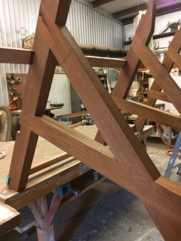 Brown wooden table frame, assembled in a workshop setting, angled view. The frame has a geometric design, and some parts are resting on a table.