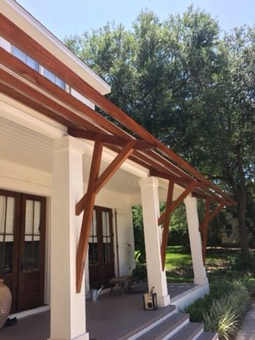 Wooden pergola over a porch with white columns and brown doors, set against a backdrop of trees and a clear sky.