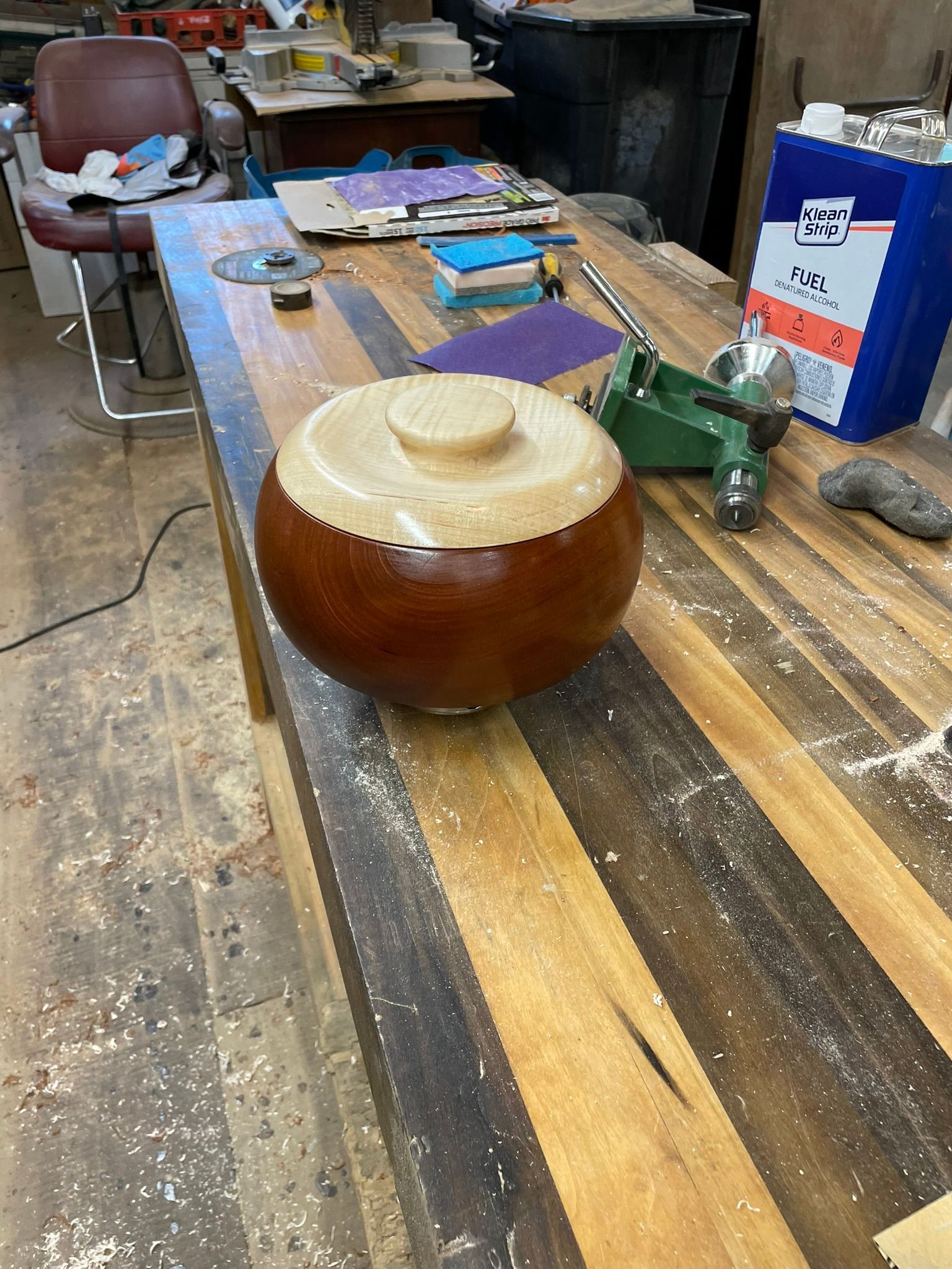 A wooden lidded bowl on a woodworking bench. The bowl is brown and cream-colored, surrounded by tools and materials.