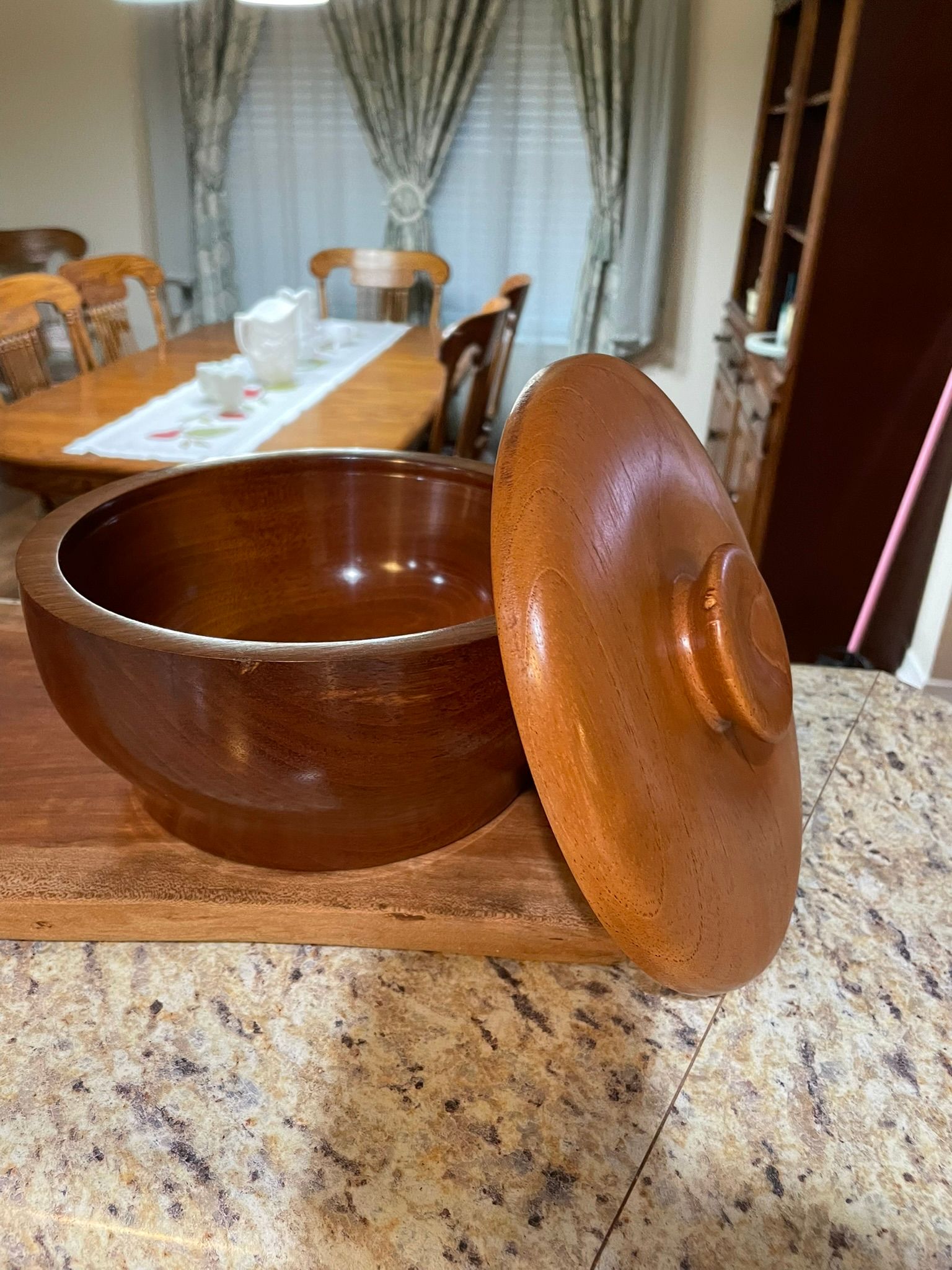 Brown ceramic bowl with lid sits on a wooden surface, with a dining room in the blurred background.