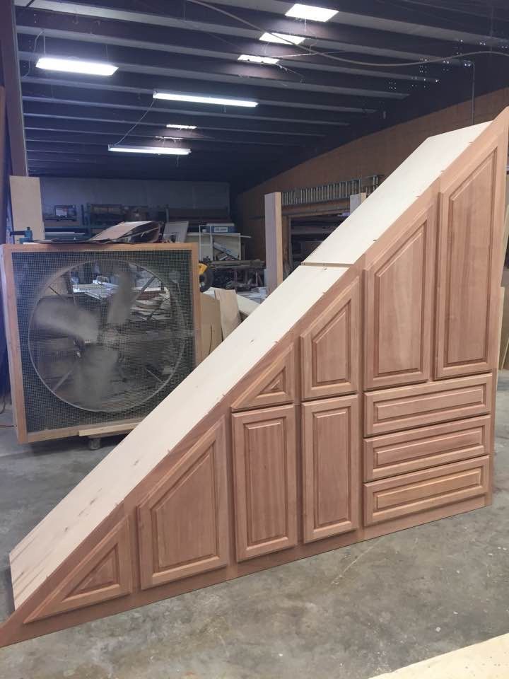 Wooden cabinet unit, slanted and against a workshop background. It has various door and drawer configurations, a light wood top, and a reddish-brown finish.