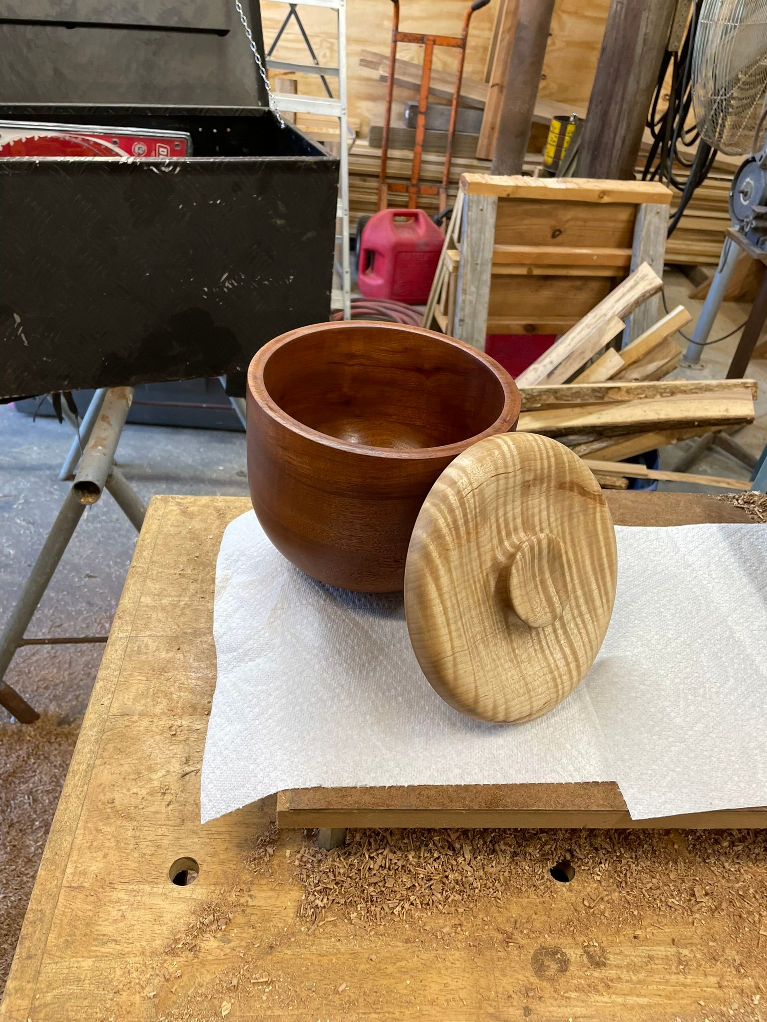 A wooden bowl with a lid on a workbench, surrounded by workshop tools and materials. The bowl is dark brown, the lid lighter.