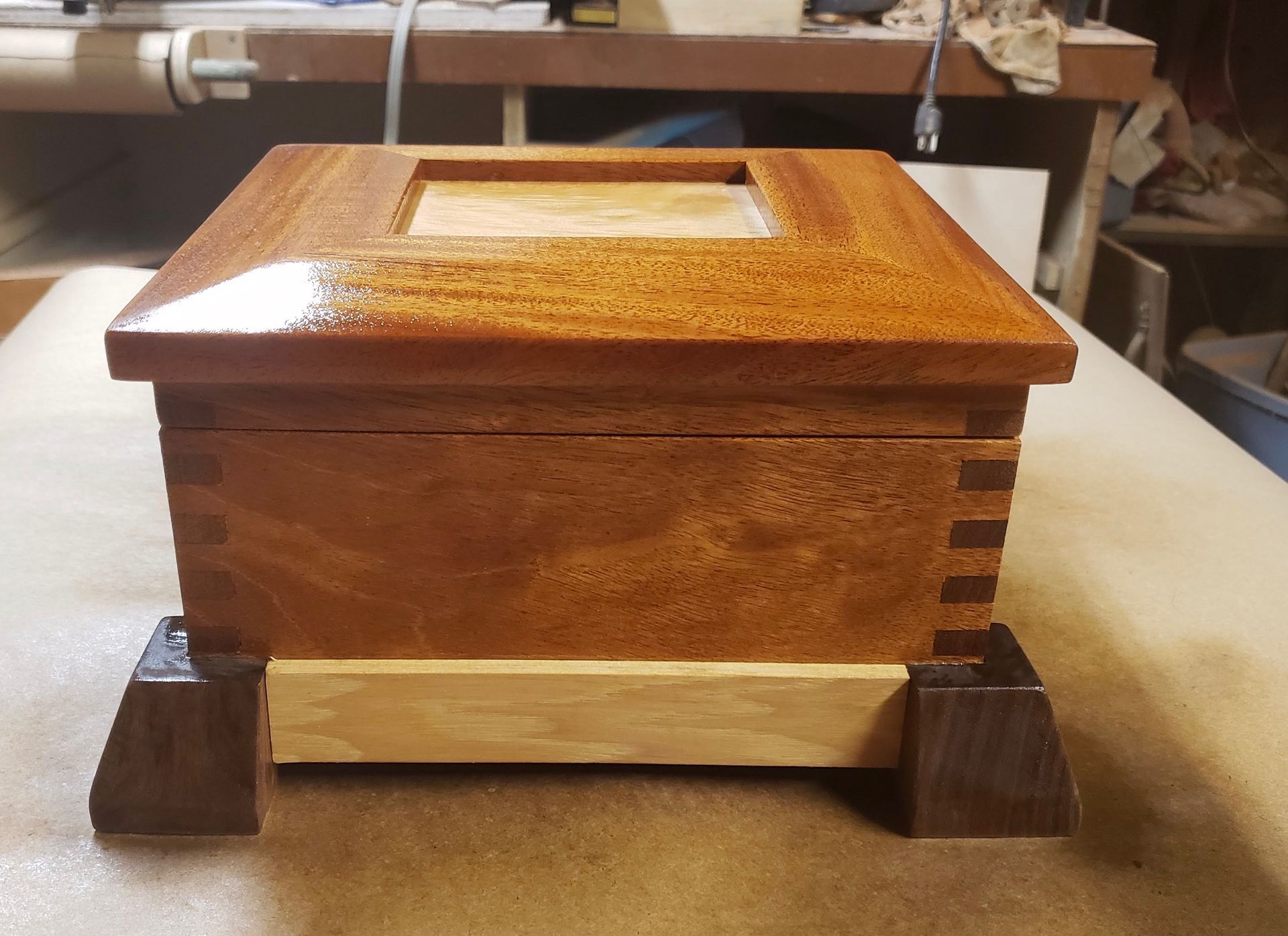 A wooden box with a hinged lid and a square cutout on top, sitting on a light-colored base with dark wooden feet.