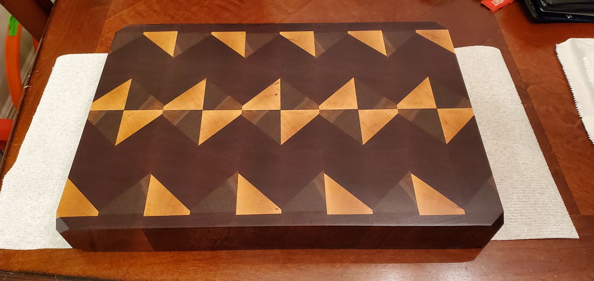 Dark wooden cutting board with zigzag pattern of dark and light wood triangles, resting on a wooden table with white towels.