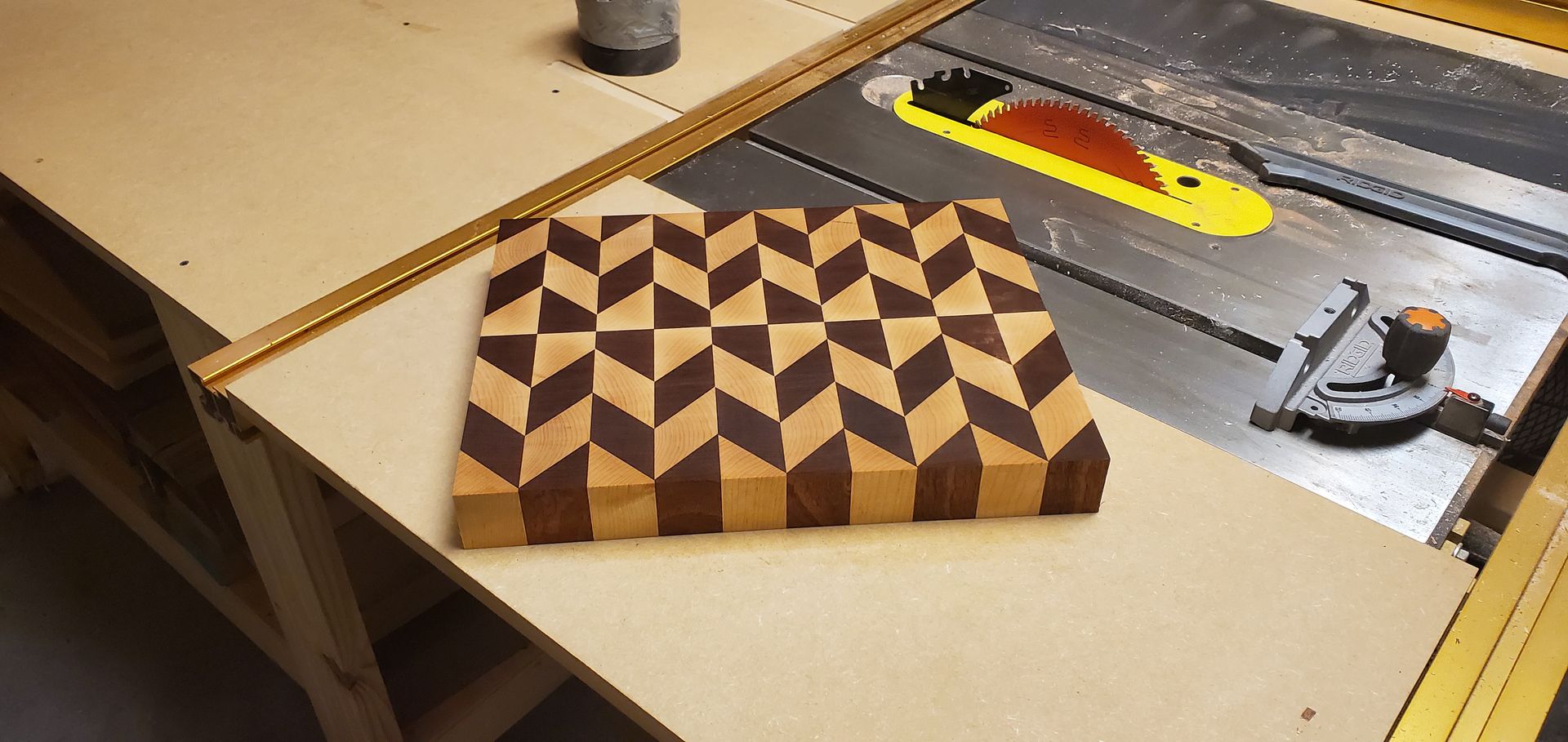 A wooden cutting board with a geometric pattern sits on a table saw. The pattern alternates dark and light wood.
