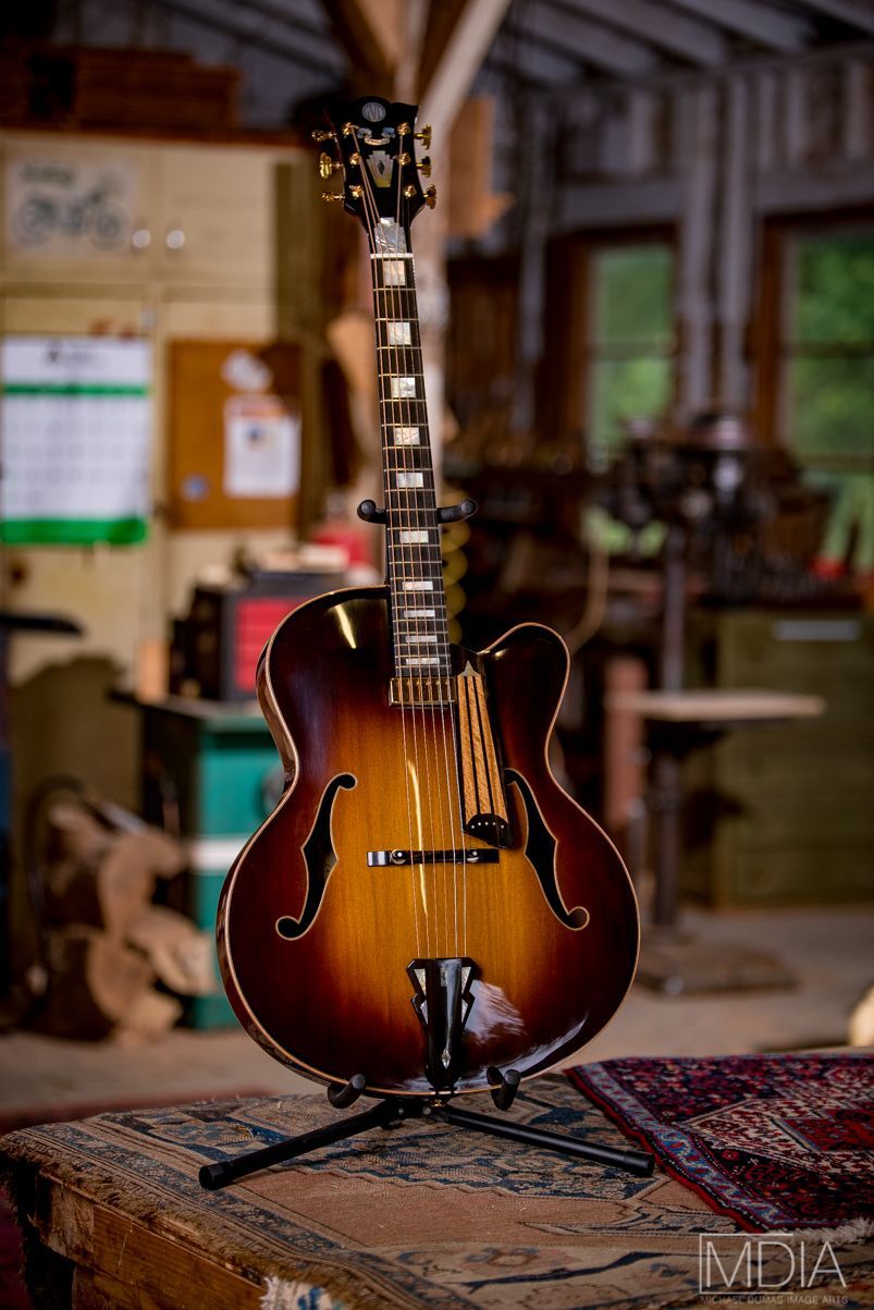 Sunburst acoustic archtop guitar on a stand in a workshop. Brown, amber, and dark hues, with intricate inlays and f-holes.
