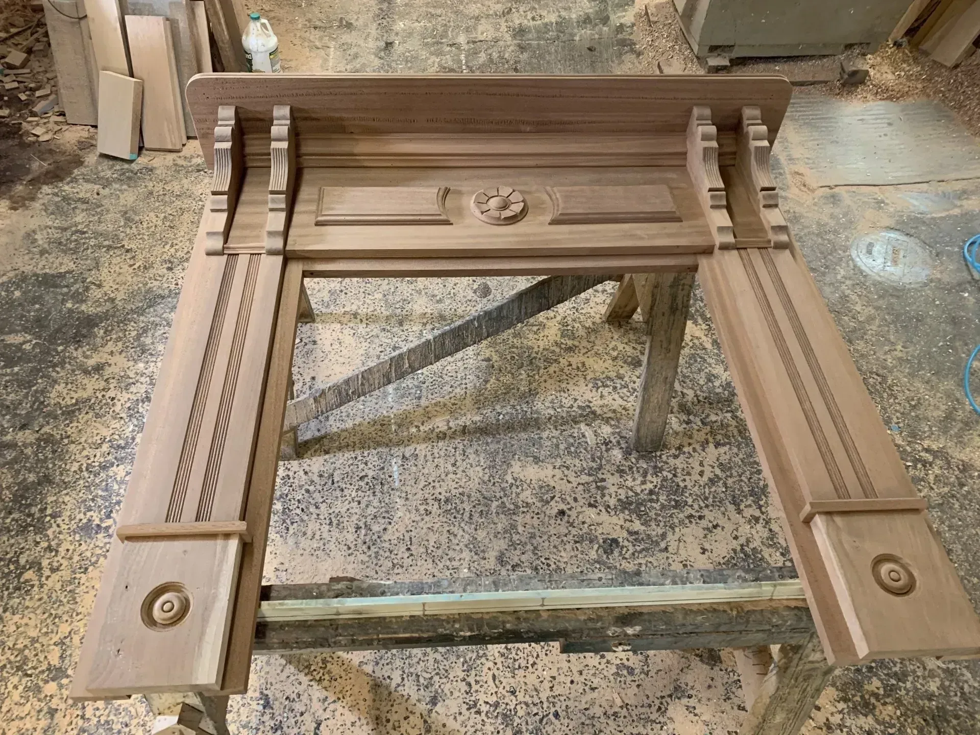 A wooden fireplace mantel under construction in a workshop. It features detailed carvings and sits on a wooden sawhorse.
