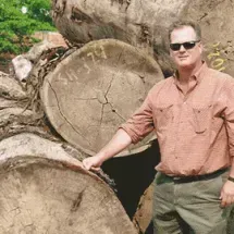 Man in sunglasses and a button-down shirt standing next to a pile of large, cut tree logs.