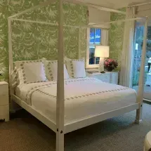 White canopy bed in a bedroom with green floral wallpaper. Includes white bedding, nightstands, and a lamp.
