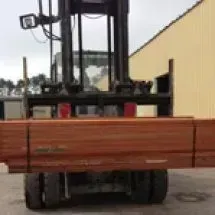 Forklift carrying a load of reddish-brown lumber outside a tan building on a cloudy day.