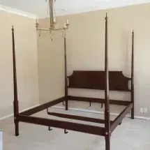 A dark wooden four-poster bed frame in an empty room with beige walls, a chandelier, and light carpeting.