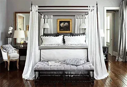 A luxurious bedroom featuring a four-poster bed with white curtains, a gray bench, and elegant decor.