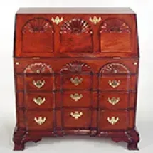 A mahogany secretary desk with fan-shaped carvings and brass hardware; it has a slanted top and several drawers.