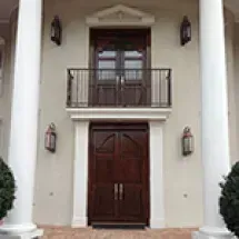 Exterior of a house with a dark wood double door, a balcony above, and white columns on either side; sconces flank the doors.
