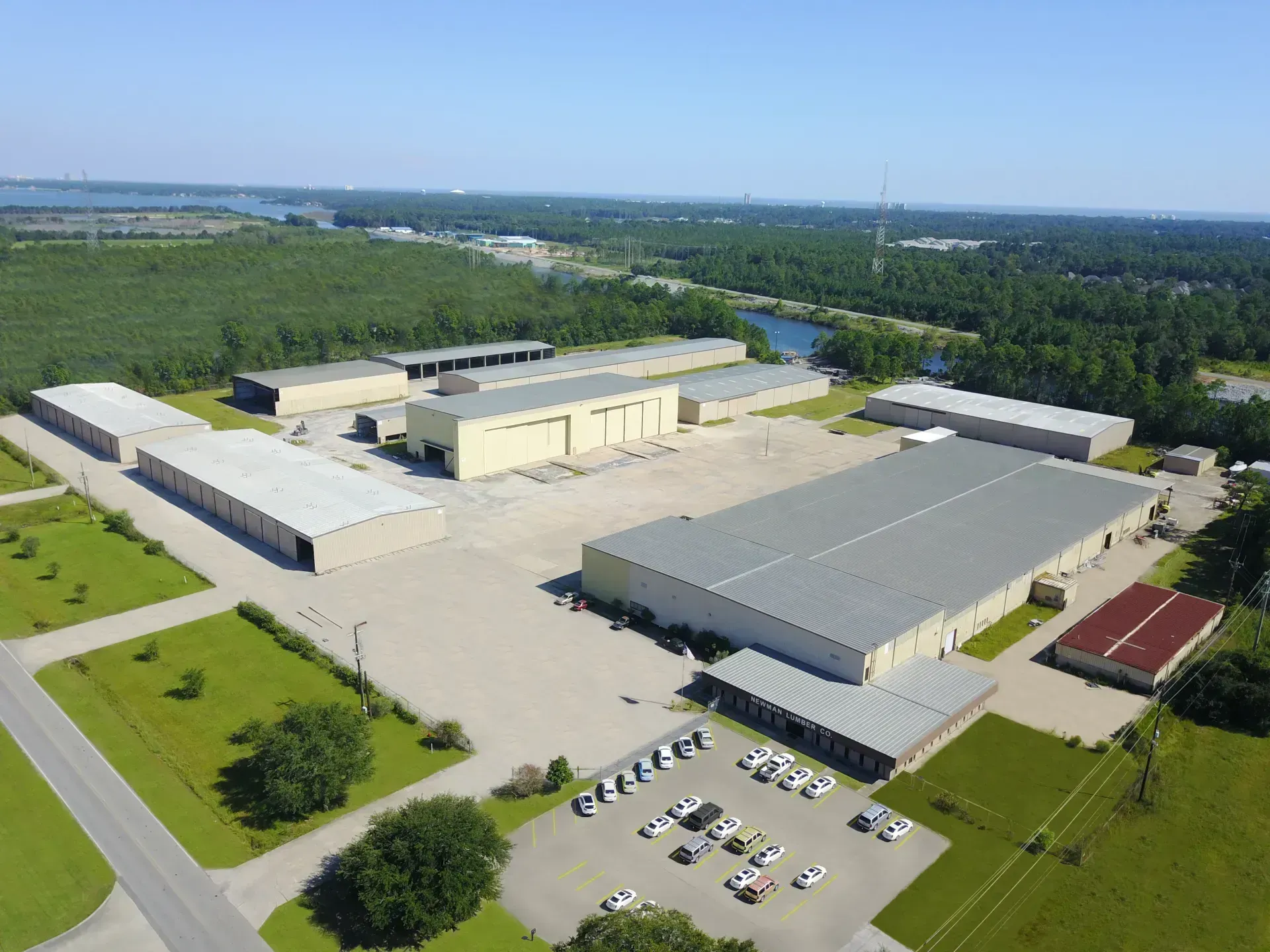 Aerial view of a large industrial complex with multiple tan buildings and a parking area surrounded by green spaces and trees.