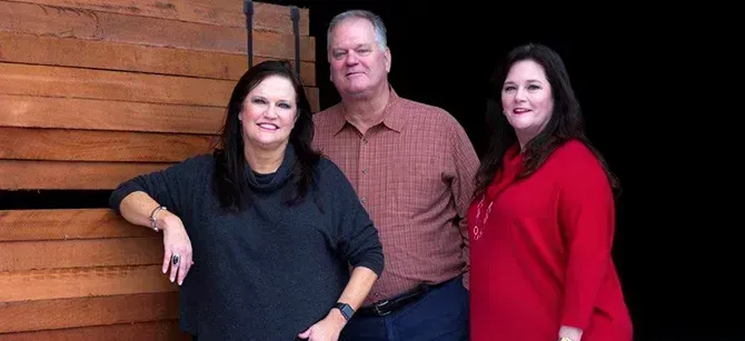 Three people posing together. A man in a button-down shirt stands between two women, one in a dark gray shirt and the other in a red sweater. Wooden wall in the background.