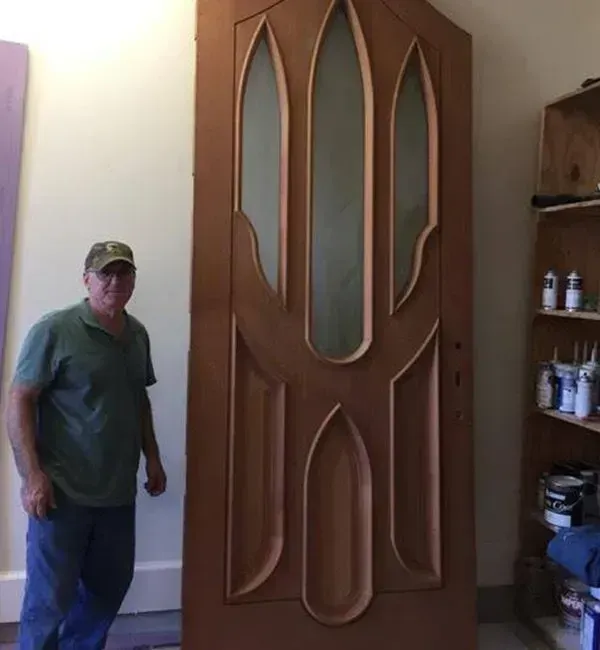Man stands next to a large wooden door with an intricate design featuring arched sections and glass panels, indoors.