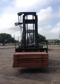 Forklift carrying a stack of reddish-brown lumber outdoors on an asphalt surface.