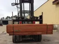 Forklift carrying a bundle of reddish-brown lumber outside a building with yellow siding.