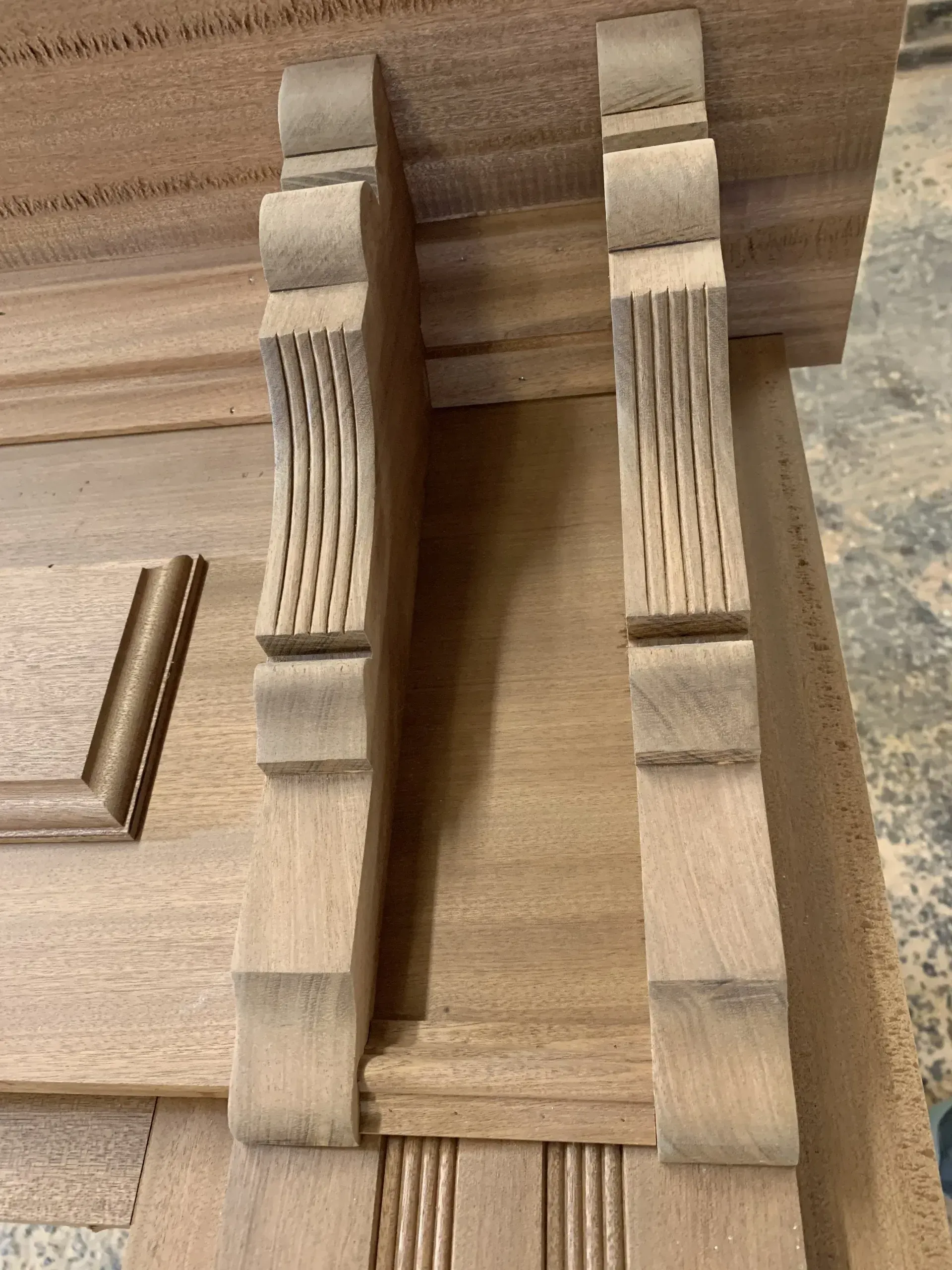Two ornate wooden corbels, freshly cut, sit on a larger wooden panel. The corbels are unfinished with carved vertical lines.