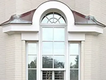 White-framed window with a rounded top, flanked by two smaller windows, set in a pale brick wall. The window is topped by a decorative white and metallic structure.
