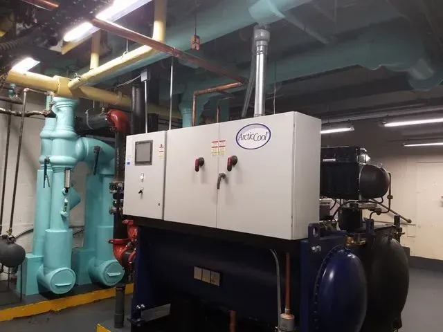 A light-colored HVAC control unit sits atop a large, blue industrial chiller in a mechanical room with overhead pipes.