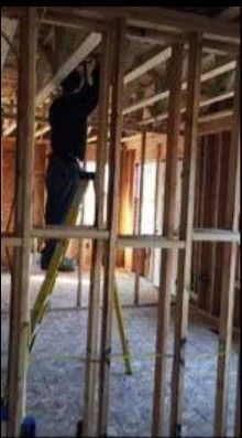 A person stands on a yellow ladder, working on the wooden framing of a room under construction.